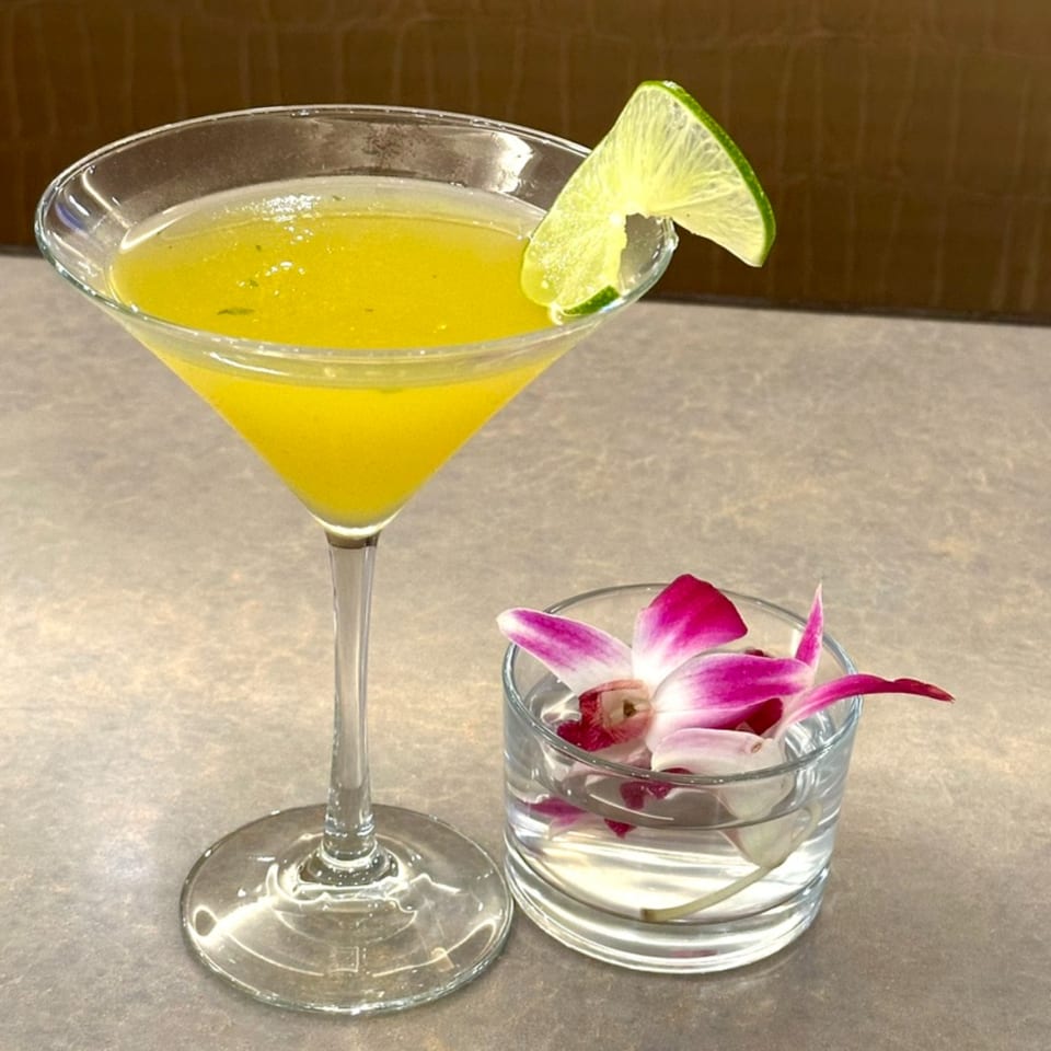 A vibrant yellow martini with a lime wedge, next to a glass with a floating pink orchid.