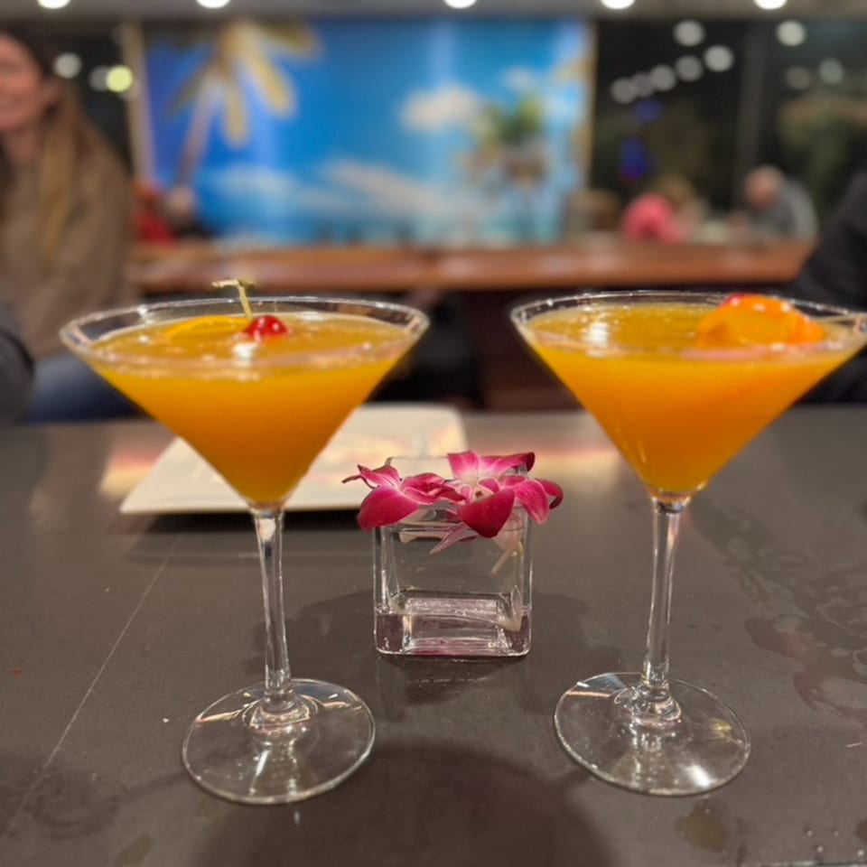 Two orange cocktails in martini glasses with fruit garnishes and a small floral centerpiece.