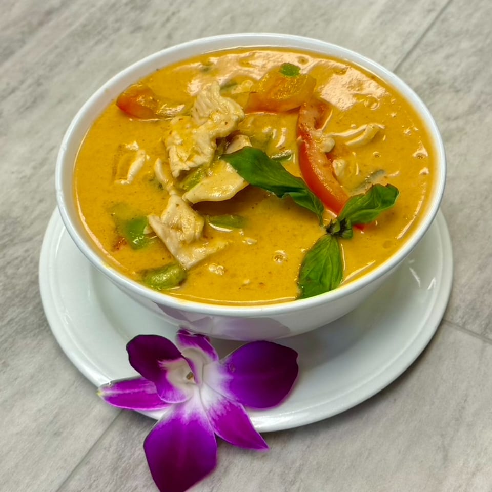 A white bowl of yellow curry with chicken, bell peppers, and basil, garnished with a purple orchid.